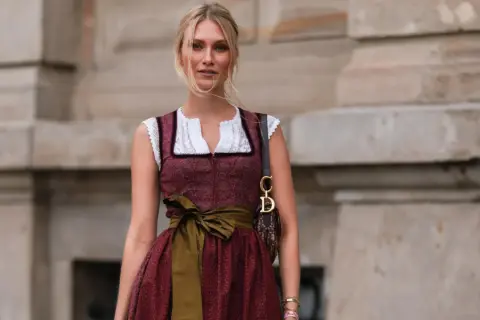 Blonde Frau in einem roten Dirndl mit weißer Bluse und einer grünen Schürze. Sie trägt eine Dior-Tasche.