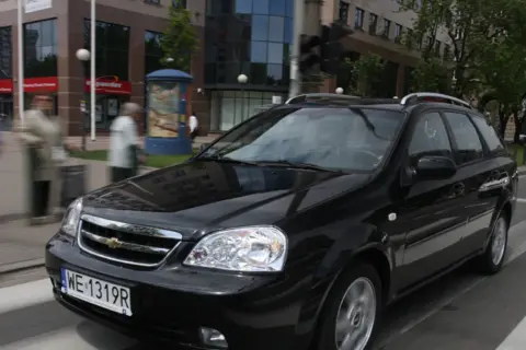 Czarny Chevrolet Lacetti kombi jedzie ulicą, mijając budynki i przechodniów.