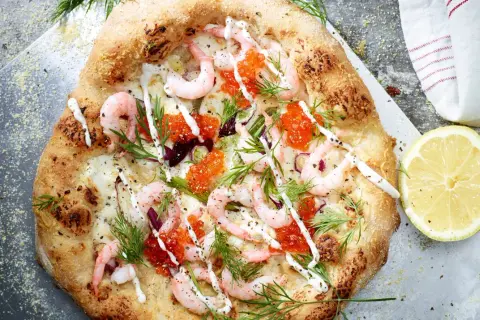 En läcker pizza med räkor och mozzarella, toppad med rom och dill. En citronskiva ligger bredvid.