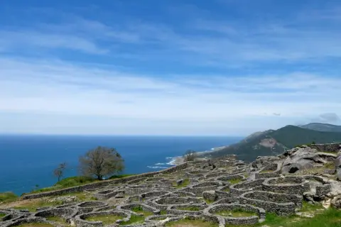 Ruinas circulares de piedra en el mirador Santa Tecla, Galicia, con el mar azul y montañas verdes al fondo.