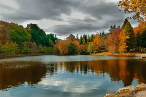 Reflejos otoñales en el lago Castiñeiras, con árboles de colores vibrantes y un cielo nublado.