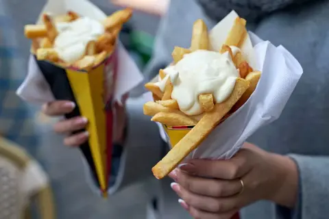 Dwie porcje frytek w papierowych rożkach, polane gęstym, białym sosem.