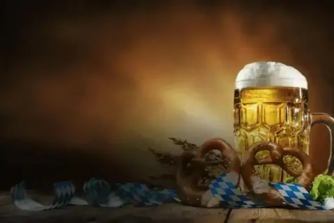 Boccale di birra dorata con schiuma abbondante, brezel e nastri a scacchi bianchi e blu, perfetti per le birre dell'Oktoberfest.