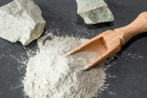 Feiner Pulver aus Magnesium-Aluminium-Silikat wird mit einer Holzschaufel auf dunklem Stein verteilt, daneben liegen zwei Brocken des Materials.