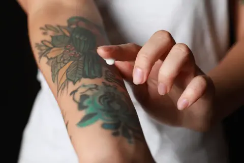 Dłoń nakłada krem na świeży tatuaż, dbając o jego prawidłowe etapy gojenia. Kolorowy wzór na ramieniu jest w trakcie regeneracji.
