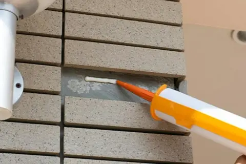 Aplikator z pomarańczową dyszą aplikuje biały silikon w szczelinę między płytkami. Dowiedz się, jak wyrównać silikon dla idealnego efektu.