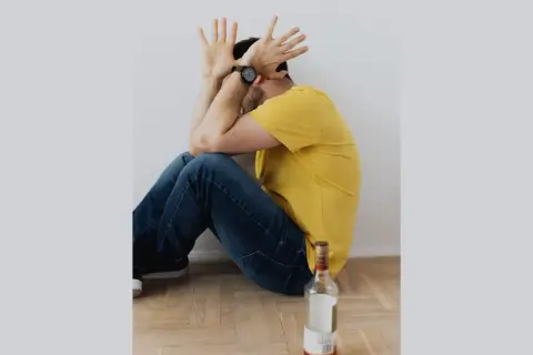 Mężczyzna w żółtej koszulce siedzi na podłodze, zakrywając twarz dłońmi. Obok leży pusta butelka. Symbolizuje trudny proces odstawienia alkoholu.
