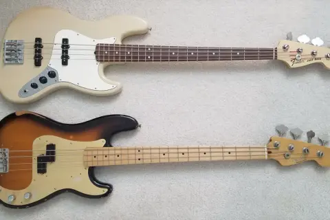 Zwei E-Bässe liegen nebeneinander: oben ein cremefarbener Jazz Bass, unten ein abgenutzter Sunburst Precision Bass.