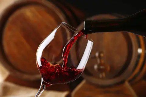 Czerwone wino leje się do kieliszka, a w tle widać drewniane beczki, które opowiadają historię wina.