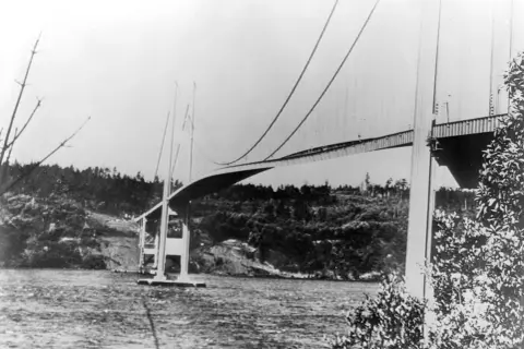 Die Tacoma-Brücke überspannt ein Gewässer, umgeben von bewaldeten Hügeln.