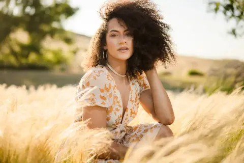 A woman with curly hair poses in a field of tall grass, her dress catching the golden hour light. This image demonstrates how to shoot with a flash for a soft, natural look.