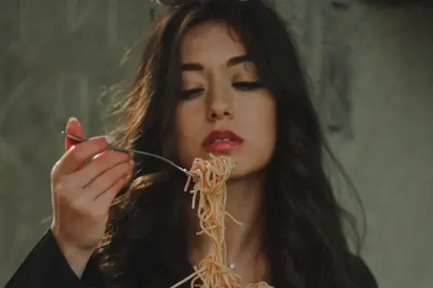 Włoska uroda w pełnej krasie. Kobieta z ciemnymi włosami i czerwoną szminką na ustach je spaghetti.