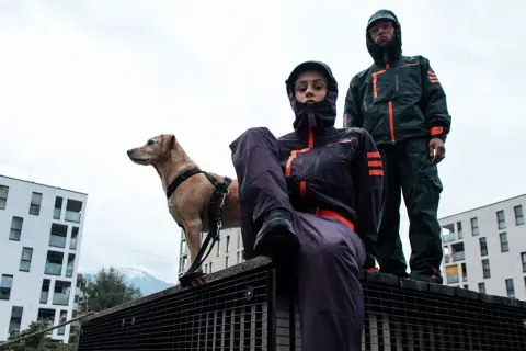 Zwei Personen in Gorpcore-Outfits und ein Hund stehen auf einem Metallgitter vor modernen Gebäuden.