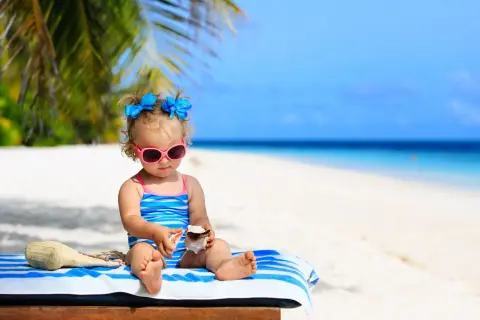Mała dziewczynka w okularach przeciwsłonecznych i stroju kąpielowym bawi się muszlami na plaży. To idealne wakacje z niemowlakiem!