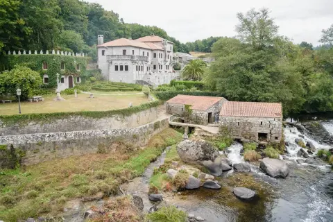 Antigua barcala con molinos de piedra junto a un río caudaloso, con una casa señorial cubierta de hiedra al fondo.
