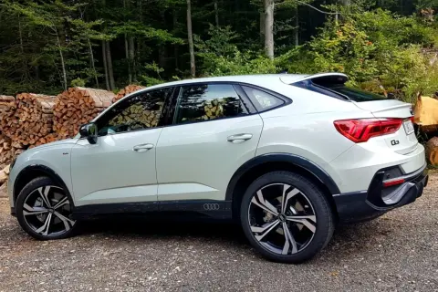 Nowy Audi Q3 2025 w kolorze białym, zaparkowany na leśnej drodze, z widocznymi stosami drewna w tle.