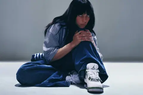Billie Eilish w luźnych dżinsach i koszuli, zamyślona, w butach Jordan. Tekst "What was I made for?" pasuje do jej nastroju.