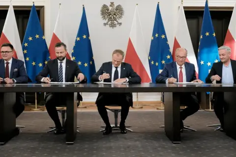 Pięciu mężczyzn w garniturach siedzi przy stole, podpisując dokumenty. W tle flagi Polski i UE. Może to być ważny moment dla sejmowej komisji śledczej.