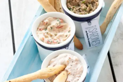 Trois ramequins de rillettes de la mer, dont une tartinade de sardines, présentés sur un plateau bleu avec des gressins.