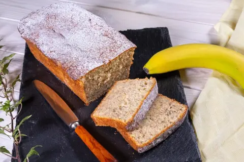 Ein saftiger Bananenkuchen, bestäubt mit Puderzucker, liegt auf einem schwarzen Brett. Daneben zwei Scheiben und eine reife Banane. Das ist der beste Bananenkuchen der Welt!