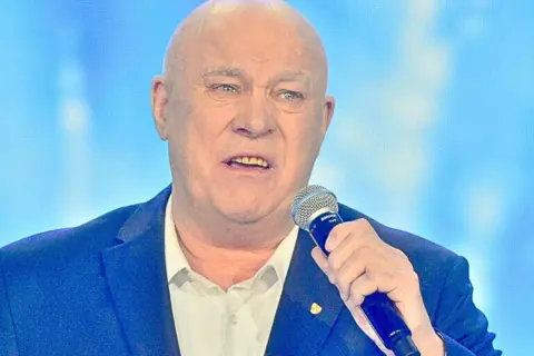 Łysy mężczyzna w garniturze trzyma mikrofon. Słychać jego głos: "Vox szczęśliwej drogi już czas!".