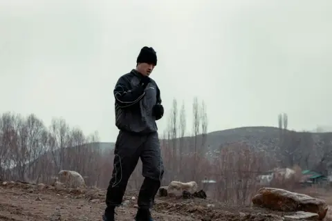 A boxer in a black tracksuit and beanie is running outdoors, practicing his footwork. The barren landscape suggests a cold day for this intense training.