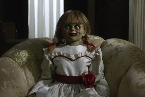 Den läskiga riktiga Annabelle dockan sitter i en blommig soffa, med sina stora ögon och röda rosett.