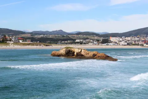 Olas rompen en la playa de Barreiros, con una roca en primer plano y el pueblo al fondo.