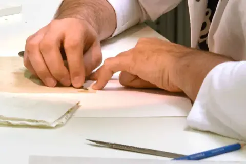 Main tenant avec précision un scalpel pour enlever de la colle sur un document ancien. Des outils et du papier sont disposés sur la table.