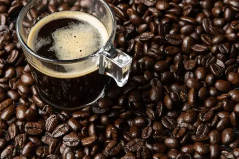 Una taza de café oscuro con espuma, rodeada de granos de café tostados. Dicen que el café es afrodisiaco, ¿será cierto?