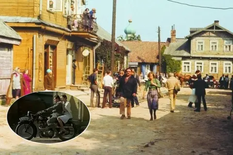 Scena z filmu "Znachor" kręcona w malowniczej wsi. W tle drewniane domy, a na pierwszym planie para na motocyklu.