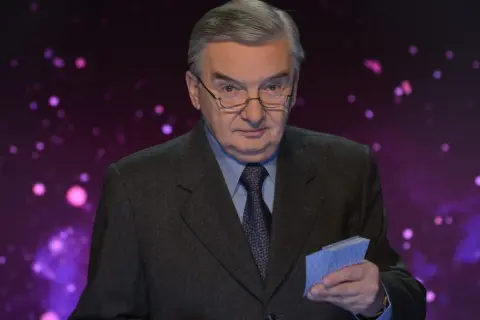 Tadeusz Sznuk w garniturze i okularach trzyma w ręku niebieskie karty na tle fioletowej, rozmytej scenografii.