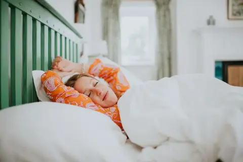 A woman in an orange patterned shirt stretches in bed, cozy under white sheets. She looks like she's getting just the right amount of light sleep.