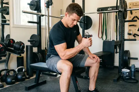 Man in a home gym using hand grippers for forearms. Weights, resistance bands, and a squat rack are visible.