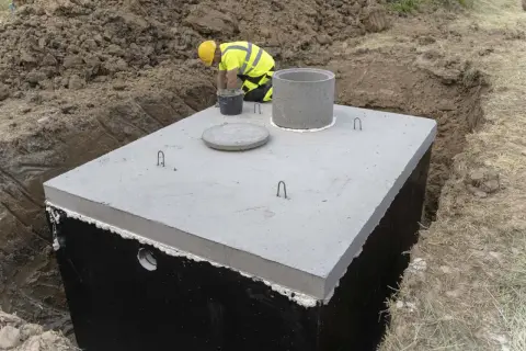 Budowa betonowego zbiornika na deszczówkę. Pracownik w żółtym kasku i kamizelce odblaskowej nadzoruje montaż.