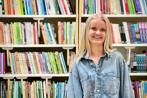 Evelina Persson ler glatt framför bokhyllor fyllda med färgglada böcker.