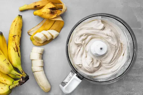 En matberedare fylld med krämig bananfrukt-glass bredvid skalade och skivade bananer.