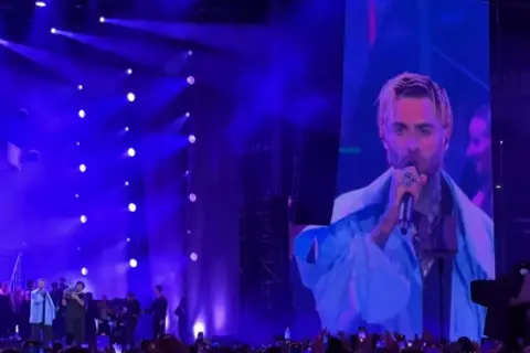 Quebonafide koncert na żywo. Artysta śpiewa do mikrofonu, a jego twarz widać na wielkim ekranie. Tłum szaleje.