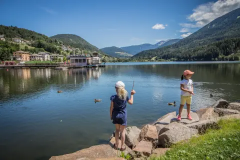 Bambine al lago di Serraia, con montagne verdi e un villaggio sullo sfondo.