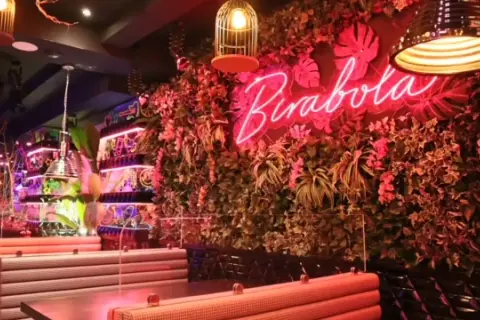 Un interior vibrante con un letrero de neón "Pirabola" sobre una pared de vegetación exuberante. Mesas y asientos de estilo cabina crean un ambiente acogedor, ideal para descubrir restaurantes originales en Zaragoza.