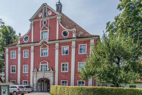Prachtvolles Gebäude des Klosters Salem mit roter Fassade, weißen Säulen und einem geschwungenen Giebel. Ein Baum spendet Schatten.