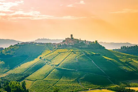Colline vitate al tramonto nelle Langhe, Roero e Monferrato, con un borgo arroccato e un castello.