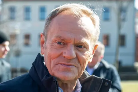 Tusk, z widocznym wiekiem, w ciemnej kurtce, na tle budynków.