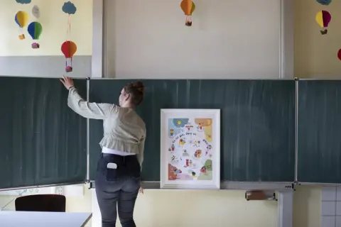 Lehrerin vor Tafel mit Poster "Es ist gut, dass es mich gibt!". Bunte Ballons hängen von der Decke. Ein Beispiel für heilende Klassenzimmer.