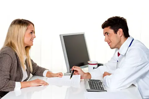 Arzt erklärt einer Frau, was passiert, wenn man die private Krankenversicherung nicht bezahlt. Sie besprechen Dokumente am Schreibtisch.