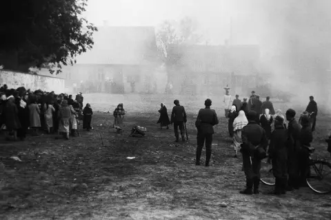 Czarno-białe zdjęcie przedstawia grupę ludzi, w tym żołnierzy, zgromadzonych na otwartym terenie przed budynkami.