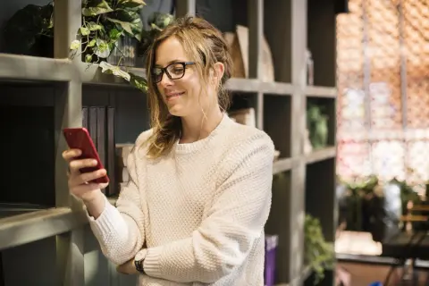 Kobieta w okularach uśmiecha się, trzymając czerwony telefon. Chyba prokrastynuję, zamiast czytać książki z półki.