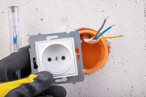 Ręka w rękawicy przygotowuje podłączenie gniazdka. Widać przewody wychodzące z puszki instalacyjnej.