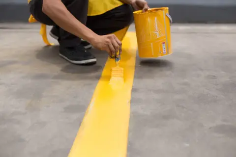Persona pinta una línea amarilla en suelo de cemento sin pulir, usando una brocha y un cubo de pintura.