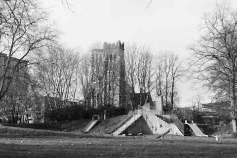 A playground with slides and stairs, captured on Ilford Pan 400 film, stands amidst bare trees with a brick tower in the background.